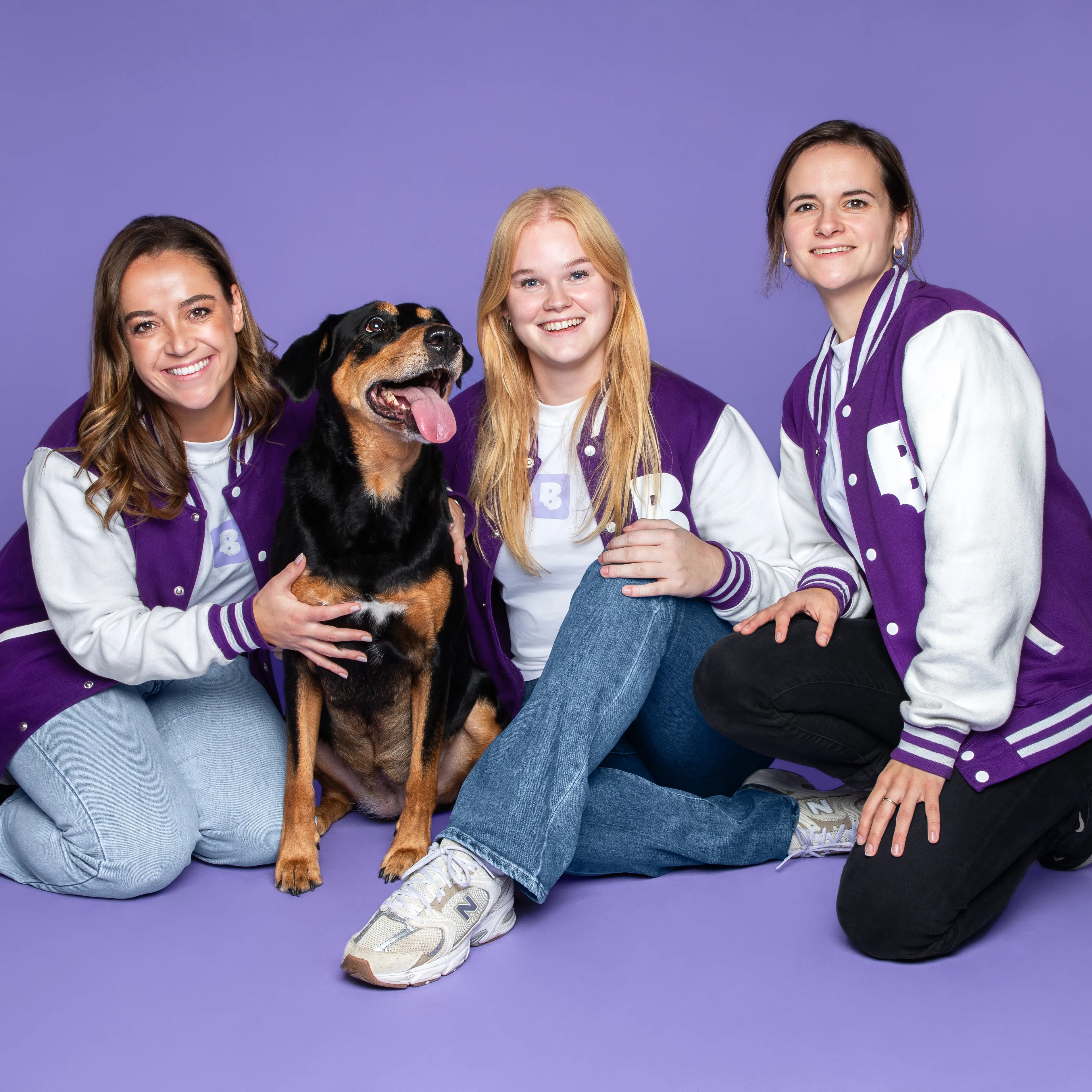 Drie vrouwen in paars-witte Braaaf-jassen poseren samen met een vrolijke zwart-bruine hond tegen een paarse achtergrond.