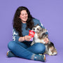 Vrouw met donker bruine krullen laat een kleine hond likken aan een Braaaf Rawhide Snacks Roll Sticks Chicken verpakking op lila achtergrond