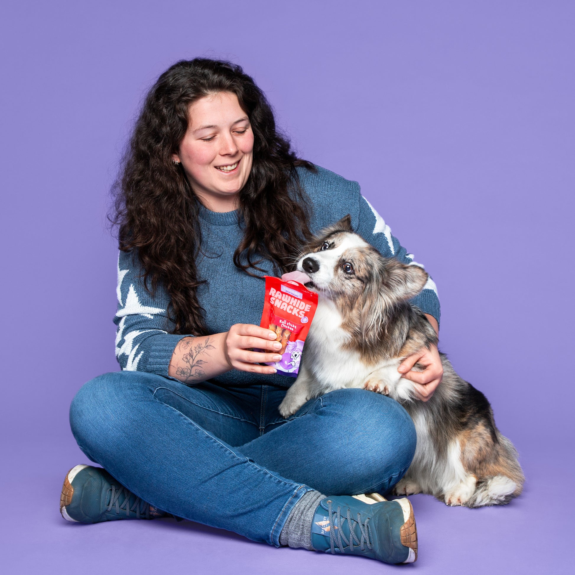 Vrouw met donker bruine krullen laat een kleine hond likken aan een Braaaf Rawhide Snacks Roll Sticks Chicken verpakking op lila achtergrond
